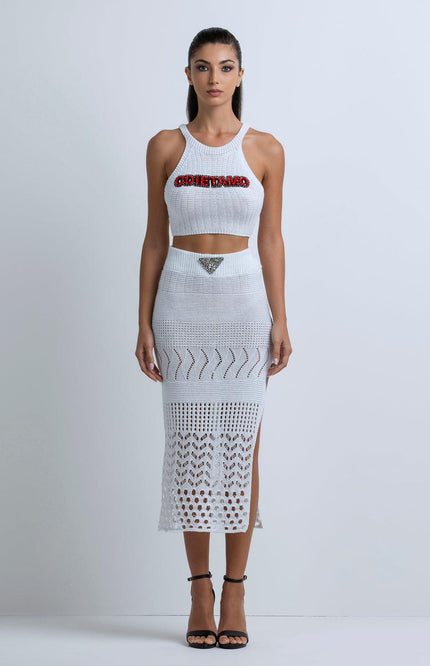 Woman wearing white crochet crop top and midi skirt with crystal accents, standing against plain background