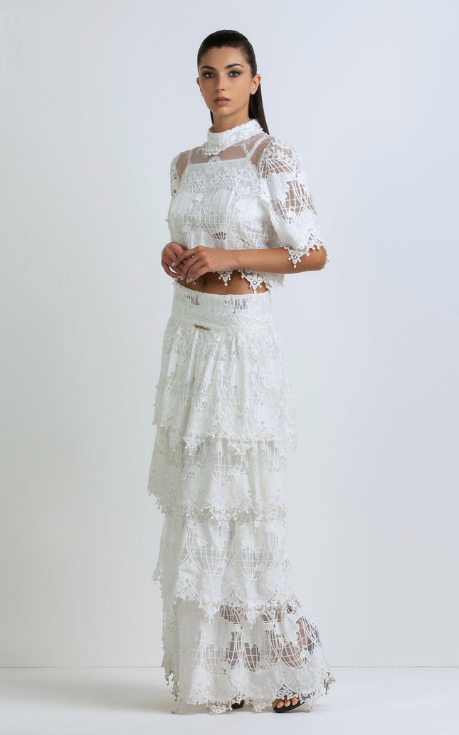 Woman in white lace two-piece dress with tiered maxi skirt and sheer details, standing indoors