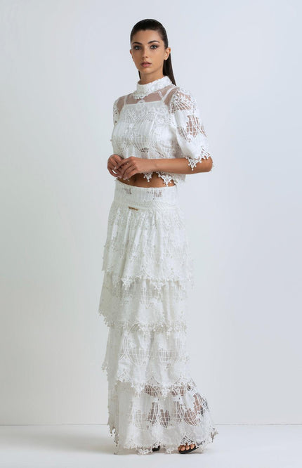 Woman in white lace two-piece dress with tiered maxi skirt and sheer details, standing indoors