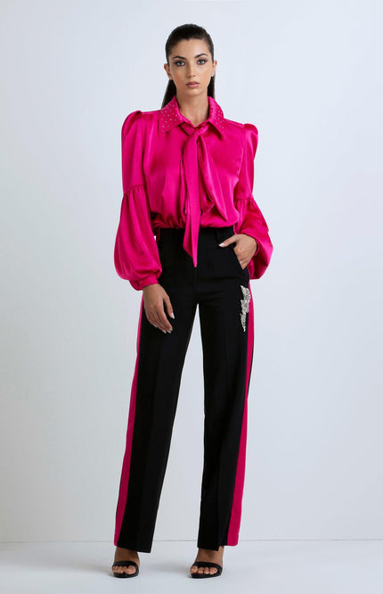 Woman wearing a bold fuchsia satin blouse with puff sleeves and black trousers with fuchsia sides and crystal embellishment, standing against a plain background.
