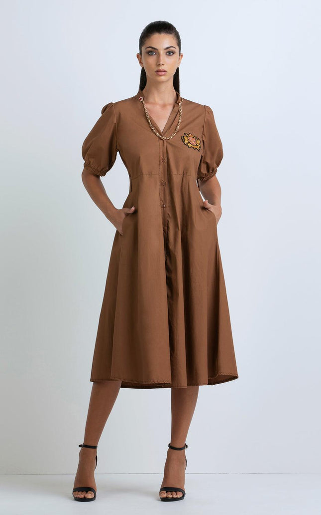 Woman wearing brown midi dress with puff sleeves, embroidered patch, gold chain detail
