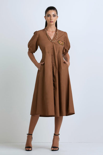 Woman wearing brown midi dress with puff sleeves, embroidered patch, gold chain detail
