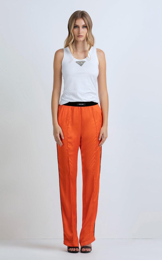 Woman wearing white tank top and bright orange satin pants with black sandals, standing against a white background