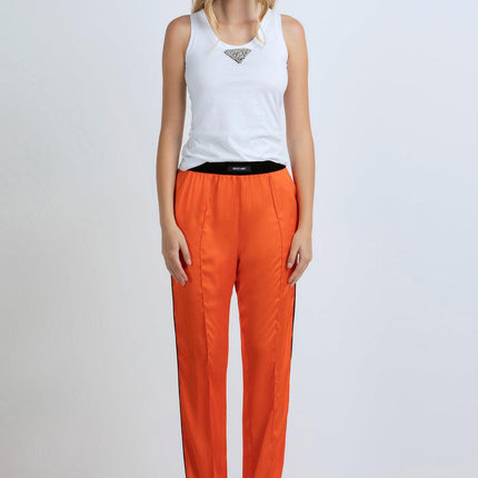 Woman wearing white tank top and bright orange satin pants with black sandals, standing against a white background