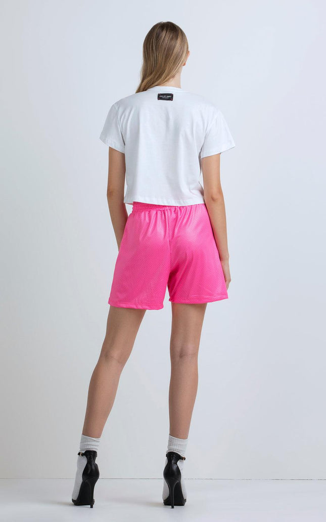 Woman in white cropped t-shirt and pink shorts, black heels, standing against plain background