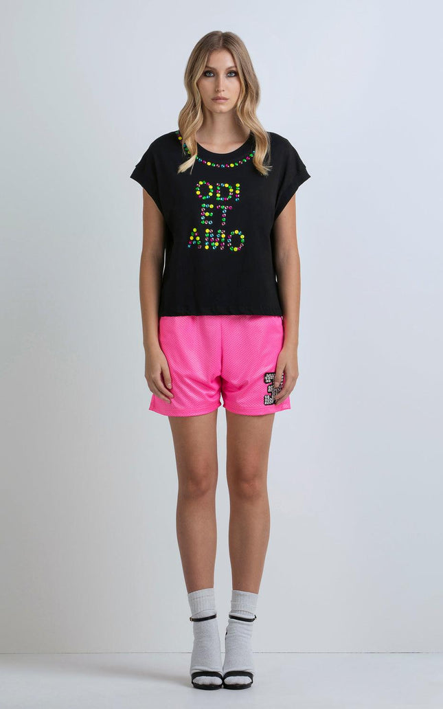 Woman in black studded t-shirt with ODI ET AMO text and bright pink shorts, standing indoors
