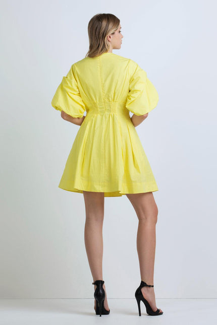 Woman in short yellow puff sleeve dress and black heels, back view, against white background