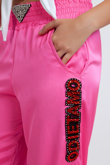 Pink satin pants with rhinestone details and ODIEETAMO lettering, hand in pocket