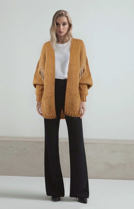 Woman wearing mustard yellow cardigan with fringe sleeves, white shirt, and black wide-leg pants