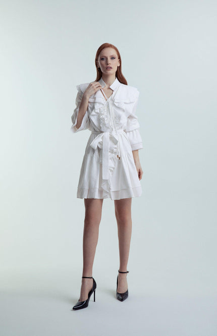 Woman wearing a stylish white belted dress with ruffled details and black heels on a plain background