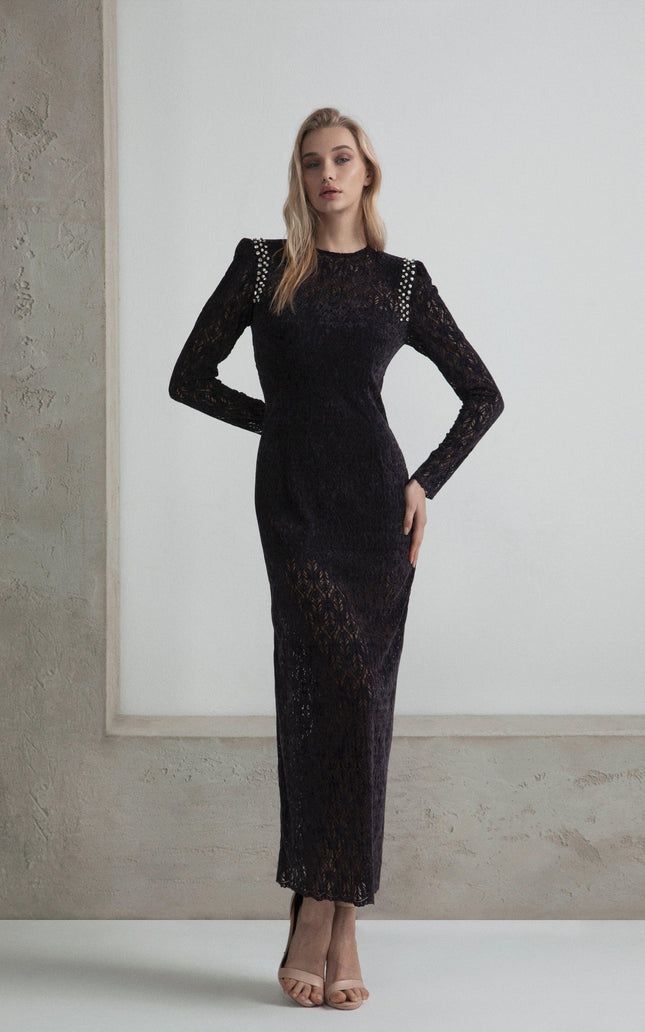 Woman in elegant black lace long dress with studded shoulders, standing against a neutral wall