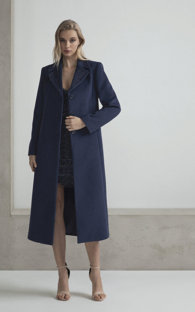 Woman in a long navy blue coat over a dark dress, standing in a minimal indoor setting