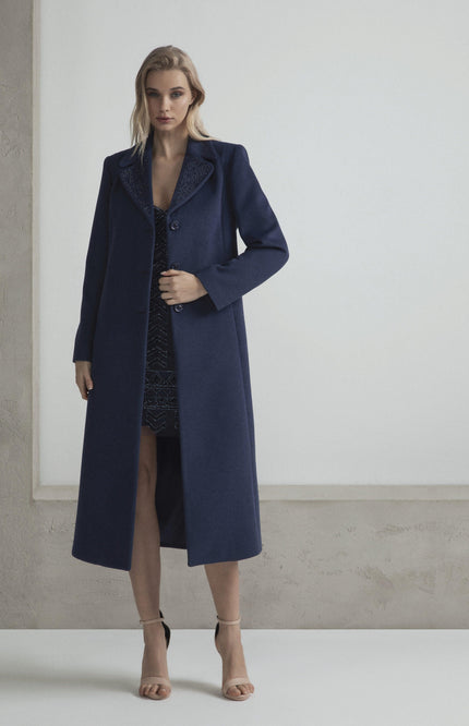 Woman in a long navy blue coat over a dark dress, standing in a minimal indoor setting