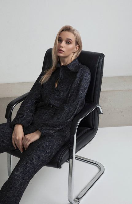 Woman in metallic dark jumpsuit sitting on modern chair, minimalist fashion look