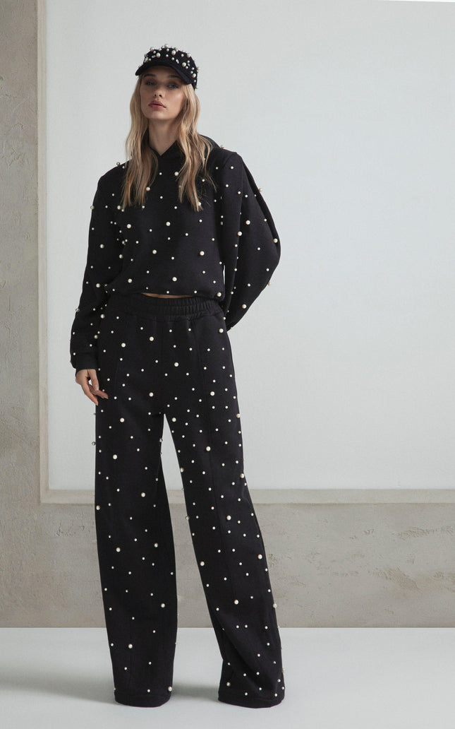 Woman in black pearl-embellished sweater, wide-leg pants, and cap, standing indoors