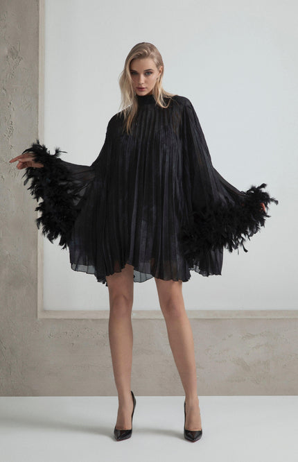 Woman in pleated black mini dress with feather trim sleeves, high heels, minimalist backdrop