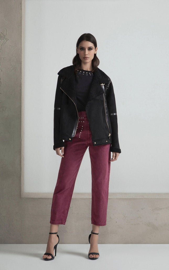 Woman in black shearling jacket, studded top, and burgundy pants, standing indoors