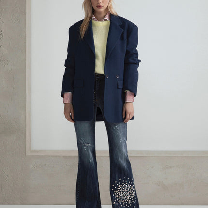Woman in oversized navy blazer, yellow sweater, and flared embellished jeans standing indoors