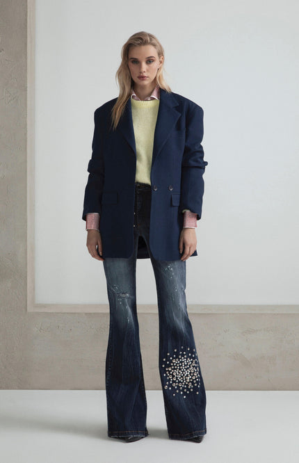 Woman in oversized navy blazer, yellow sweater, and flared embellished jeans standing indoors