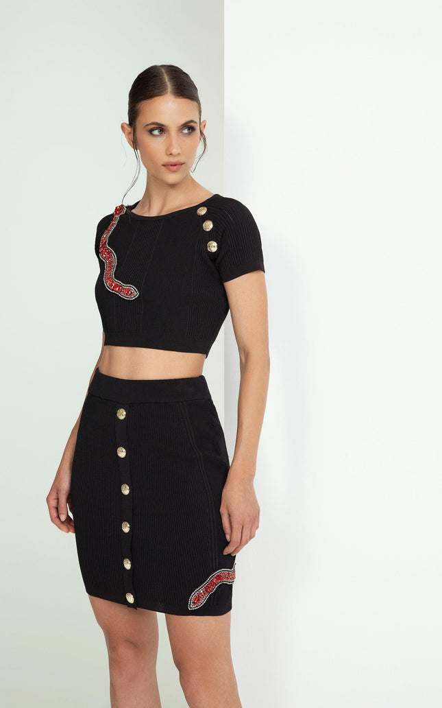 Woman wearing black crop top and skirt set with gold buttons and red snake embellishments