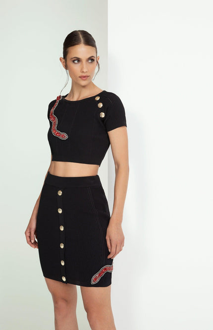 Woman wearing black crop top and skirt set with gold buttons and red snake embellishments