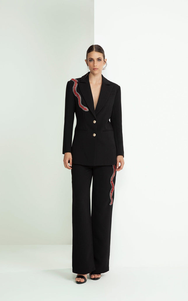Woman in a black tailored suit with red sequin snake detail, wide leg pants, minimalist setting