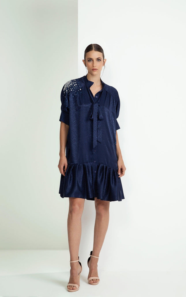 Woman wearing a navy blue short dress with embellished shoulder, ruffle hem, and bow tie neck.