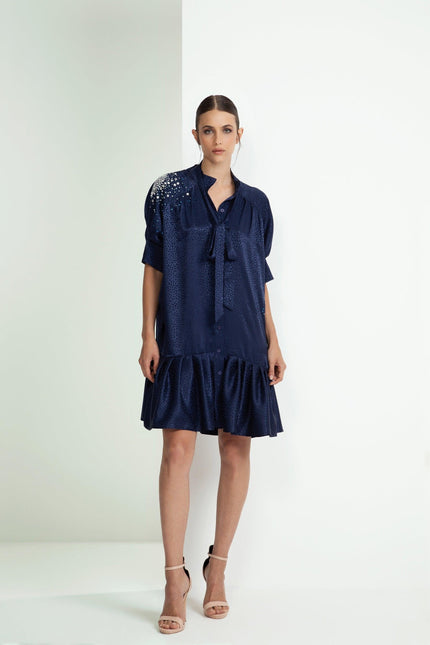 Woman wearing a navy blue short dress with embellished shoulder, ruffle hem, and bow tie neck.