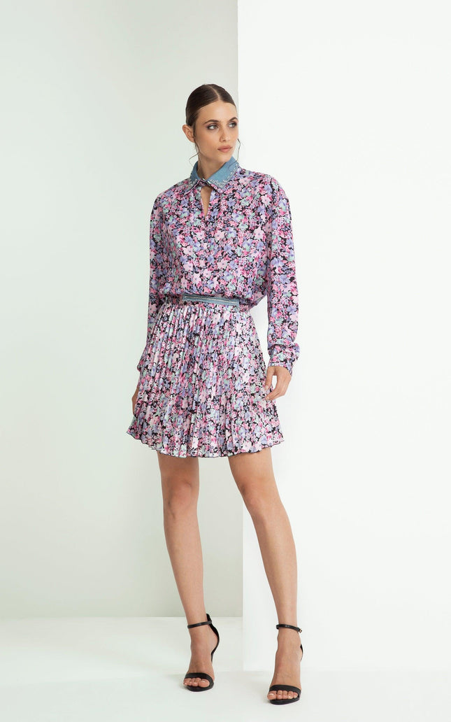 Woman wearing a floral blouse and pleated mini skirt with black heels, standing indoors