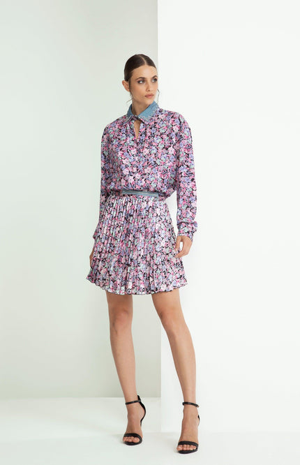 Woman wearing a floral blouse and pleated mini skirt with black heels, standing indoors