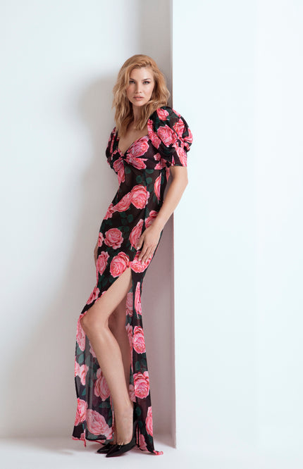 Un elegante abito lungo semi-velato in poliestere caratterizzato da una romantica stampa a rose, scollo a cuore con maniche a palloncino e un audace spacco sulla coscia.