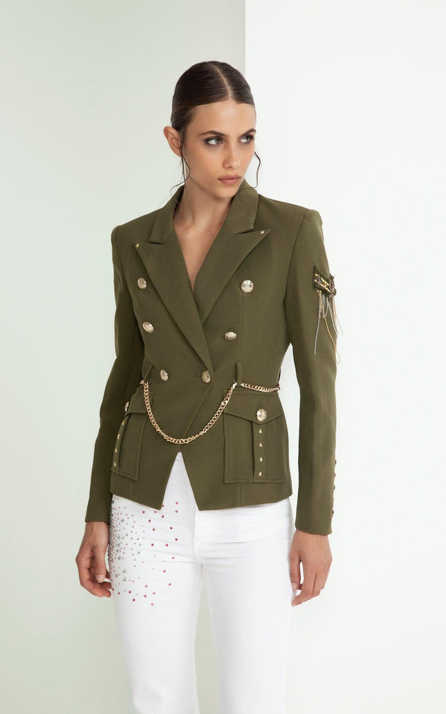 Woman wearing olive green military-style blazer with gold buttons and chain, white pants with studs