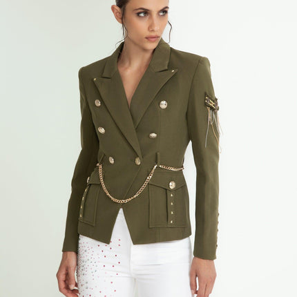 Woman wearing olive green military-style blazer with gold buttons and chain, white pants with studs