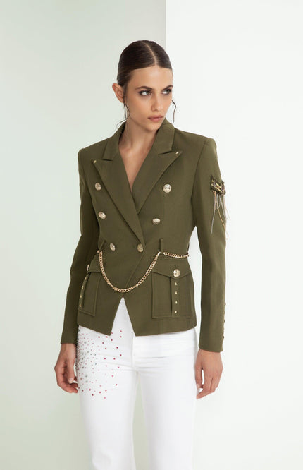 Woman wearing olive green military-style blazer with gold buttons and chain, white pants with studs