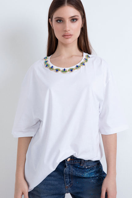 Modella che indossa la t-shirt oversize bianca con applicazione di pietre gioiello multicolor sul collo e jeans blu scuro.
