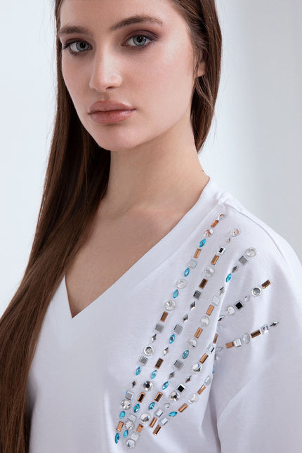 Primo piano di t-shirt donna bianca con scollo a V e applicazione di pietre gioiello colorate e strass sulla spalla