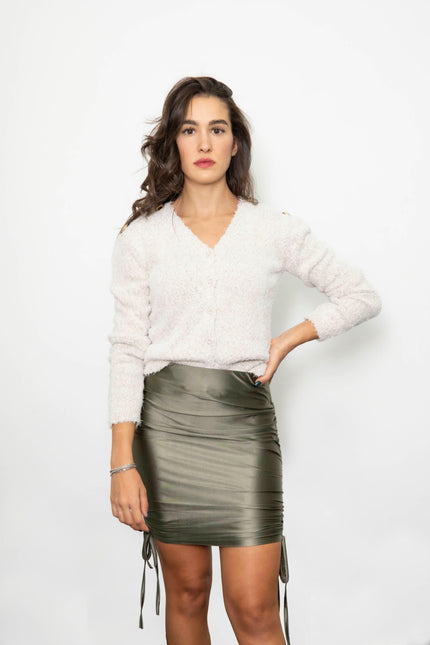 Woman in white fuzzy sweater and olive green ruched satin mini skirt, standing against white background