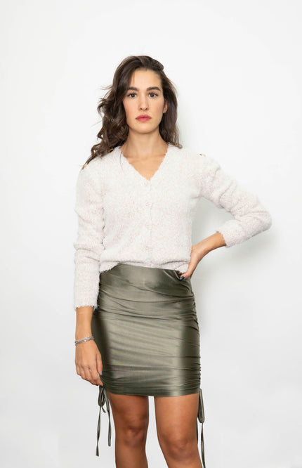 Woman in white fuzzy sweater and olive green ruched satin mini skirt, standing against white background